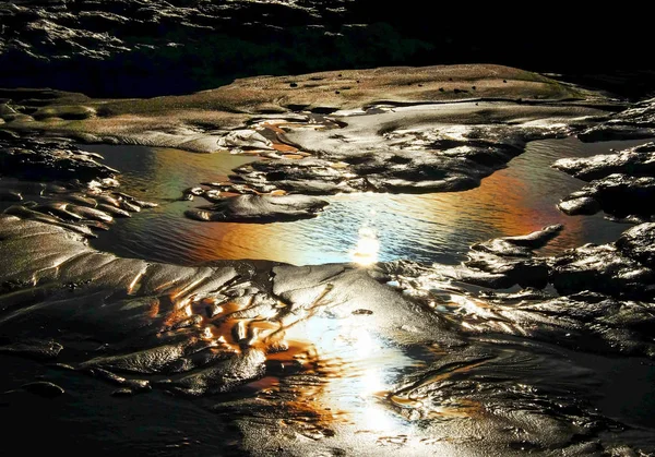 Close up of a rock pool with rock and sand around a pool of water with rainbow colours from sun light reflecting in the water, the image is back lit showing all the detail and texture in the sand and rocks, abstract and graphic resource, text space,