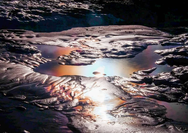 Close up of a rock pool with rock and sand around a pool of water with rainbow colours from sun light reflecting in the water, the image is back lit showing all the detail and texture in the sand and rocks, abstract and graphic resource, text space,