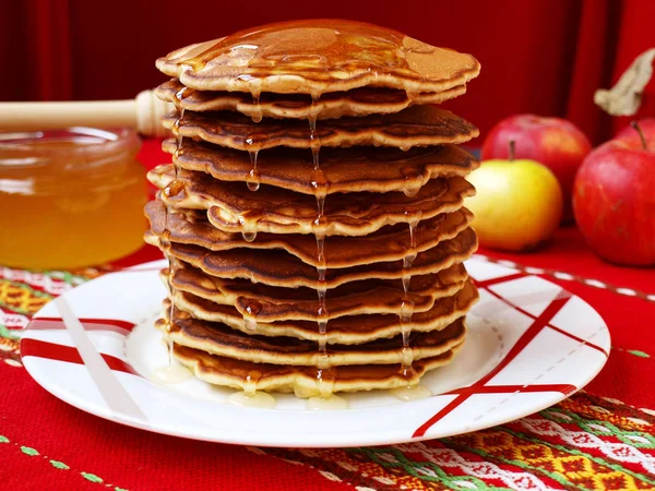 Pancakes are located on a plate. Honey drains from above. In the background, apples and honey