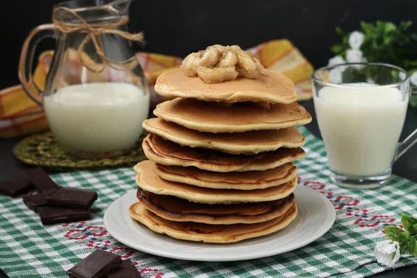 Panqueques con plátanos en un plato sobre un fondo oscuro, detrás hay una jarra y un vaso de leche — Foto de Stock