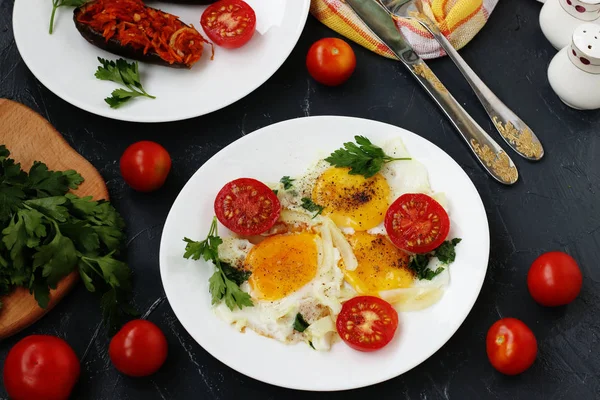 Scrambled eggs with cherry tomatoes are located on a white plate on a dark background, the photo there baked eggplants, parsley, cutlery, top view — Stock Photo, Image