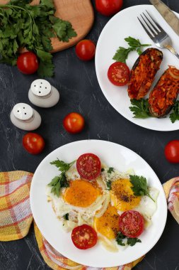 Çırpılmış yumurta ve kiraz domatesleri koyu arkaplanda beyaz bir tabakta, fırınlanmış patlıcan, maydanoz, çatal bıçak takımı.