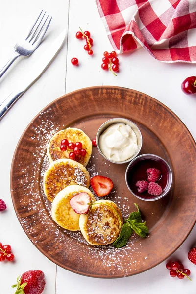 Cottage cheese pancakes, homemade traditional Ukrainian dish syrniki with berry sauce and sour cream on a brown plate on a white background, vertical photo — Stock Photo, Image