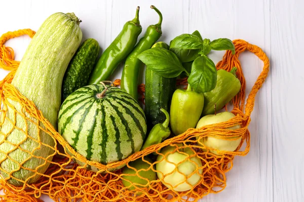Πράσινα λαχανικά και φρούτα σε πορτοκαλί επαναχρησιμοποιήσιμη τσάντα πλέγματος αγορών σε λευκό φόντο: κολοκυθάκια, αγγούρια, πιπεριά, καυτές πιπεριές, μήλα, αχλάδι, καρπούζι και βασιλικό, οριζόντιος προσανατολισμός — Φωτογραφία Αρχείου
