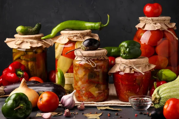 Різні мариновані овочі в скляних банках для тривалого зберігання: салат з баклажанами, перцем, огірками, помідорами та змішаними овочами на темному фоні, горизонтальна орієнтація — стокове фото