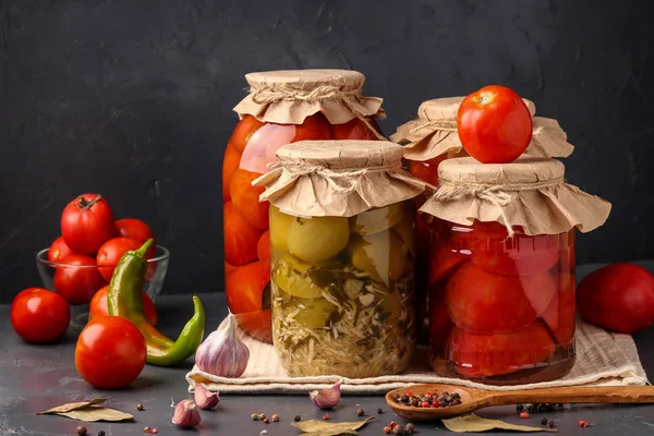 Different types of canned tomatoes, halves of tomatoes, green tomatoes with horseradish in glass jars for long-term storage on a dark background, horizontal orientation