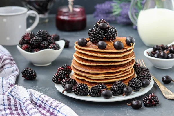 Pancakes with maple syrup, blackberries and currants are stacked on a plate, in the background berries and a jug of milk, horizontal orientation — Stock Photo, Image