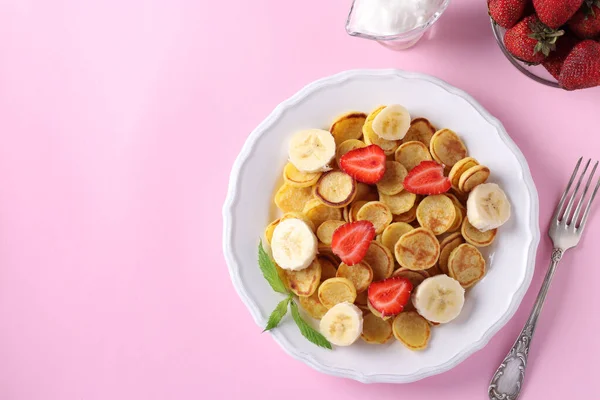 Clătite mici la modă pentru micul dejun cu căpșuni, banane și smântână în farfurie albă pe fundal roz. Vedere de sus. Copiați spațiul pentru text sau design — Fotografie, imagine de stoc