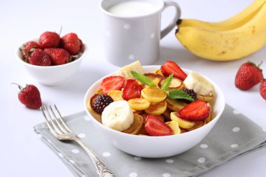 Trendy tiny pancakes for breakfast with strawberry, blackberry and banana in bowl on white background and cup milk.