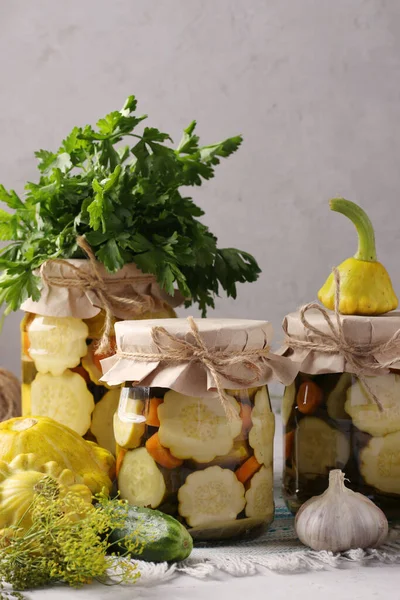 Homemade pickled patissons, cucumbers with carrots and garlic in glass jars and fresh ingredients on a light gray background, Vertical format — Stock Photo, Image