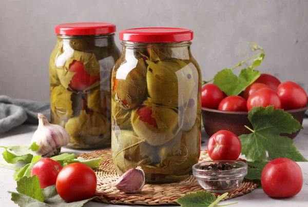 Marinated tomatoes with garlic in grape leaves in two glass jars on grey background. Closeup. Horizontal format — Stock Photo, Image
