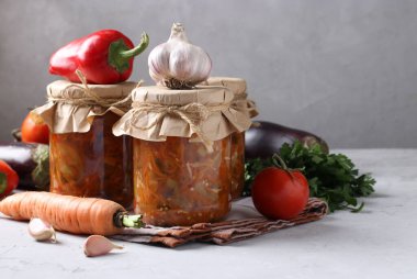 Patlıcan, havuç, biber, sarımsak ve domatesli sebze salatası gri arka planda cam kavanozlarda, yatay formatta. Metin için yer.