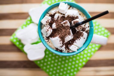 Beyaz marşmelovlu kahve, rendelenmiş çikolata, bardakta siyah bir pipet, yeşil bir peçetede, ahşap bir masada..