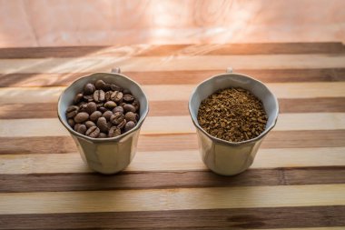 Ahşap bir tahtanın üzerinde iki fincan dolusu kavrulmuş kahve çekirdeği ve hazır kahve tozu var..