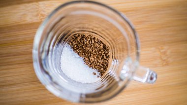 Şeffaf bir fincan ahşap bir arka planda durur, hazır kahve ve şeker bardağın dibine dökülür..