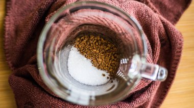 Şeffaf bir fincan ahşap bir arka planda durur, hazır kahve ve şeker bardağın dibine dökülür..