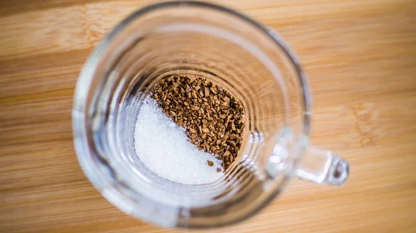 Şeffaf bir fincan ahşap bir arka planda durur, hazır kahve ve şeker bardağın dibine dökülür..