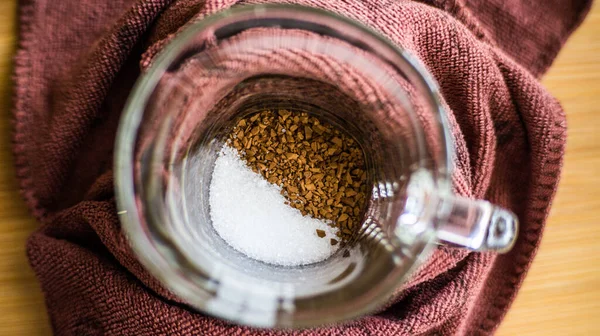 Şeffaf bir fincan ahşap bir arka planda durur, hazır kahve ve şeker bardağın dibine dökülür..