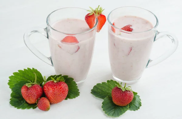 两个透明的玻璃杯 牛奶草莓鸡尾酒在白色的木桌上 旁边的草莓浆果与叶子 — 图库照片