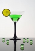 Sklenička Martini se zeleným nápojem a plátkem vápna, stojící na tmavém reflexním stolku vedle zelených průsvitných koulí