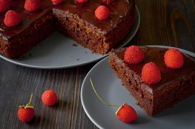 Bir parça siyah çikolatalı kek gri bir tabakta, büyük bir tabağın yanında, parlak kırmızı böğürtlenli bir kek..