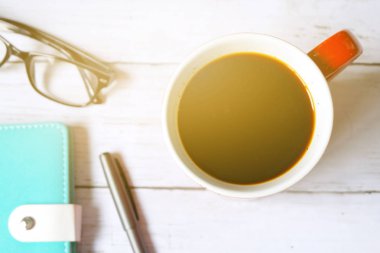 Seçici odak ve beyaz ahşap zemin üzerinde siyah kahve defter, kalem ve güneş gözlüğü ile kırmızı kupa üstten görünüm.