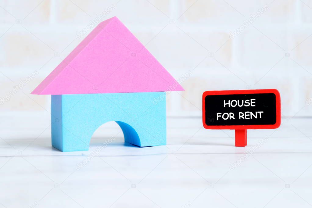 Selective focus of red blackboard written with HOUSE FOR RENT with house model on white wooden background. Real estate theme.