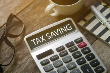 Bir fincan kahvenin, bardakların, kalemin, defterin ve hesap makinesinin seçmeli odak noktası. Tahta arka planda Vergi Tasarruf ile yazılmış..