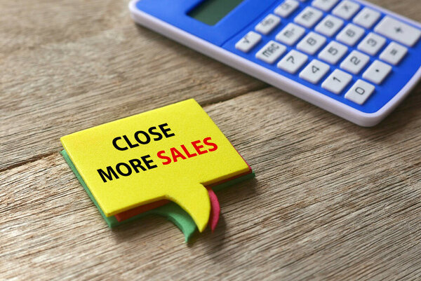 Selective focus of calculator and stack of speech bubbles written with ' Close More Sales ' on wooden background.