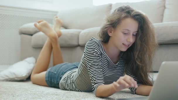 Menina adolescente bonito em um tapete funciona com laptop. Steadicam tiro de menina adolescente feliz perto de um sofá com laptop. Adolescente usa redes sociais, lê notícias, sorri e olha para a câmera . — Vídeo de Stock