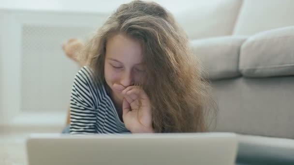 Nettes Teenie-Mädchen auf einem Teppich arbeitet mit Laptop. Steadicam Aufnahme eines glücklichen Teenie-Mädchens in der Nähe eines Sofas mit Laptop. Teenie-Mädchen nutzt soziale Netzwerke, liest Nachrichten, lächelt und blickt in die Kamera. — Stockvideo