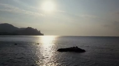 Geriye doğru deniz yüzeyi kayalar ve taşlar üzerinde güneş doğarken uçan. Dağlar, taşlar, taşlar su ve deniz dalgaları. Kırım'da Kara Dag Dağı havadan görünümü.