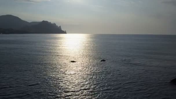 Volare all'indietro sulla superficie del mare con rocce e pietre all'alba. Montagne, rocce, pietre nell'acqua e onde del mare. Veduta aerea della montagna di Kara Dag in Crimea . — Video Stock