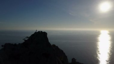 Aerial silhouette of young woman on the top of a mountain facing the sea. Flight over Lady on the summit raising up hands in beautiful scenery.