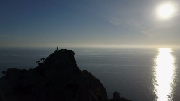 Aerial silhouette of young woman on the top of a mountain facing the sea. Flight over Lady on the summit raising up hands in beautiful scenery. — ストック動画