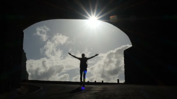 カメラは、トンネルから太陽の光に向かって歩いている女性のシルエットに従います。明るい光のバースト空中で腕を上げる. — ストック動画