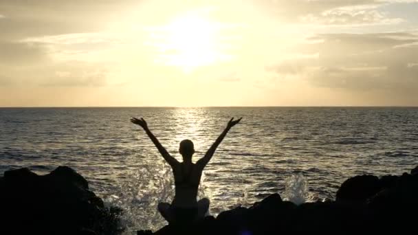 Silueta de una mujer sentada sobre rocas al atardecer observando olas y levantando brazos en el aire. Cámara lenta cinematográfica — Vídeos de Stock
