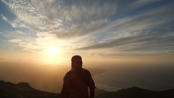 A câmera segue um homem se aproximando de um penhasco sob forte vento acima do oceano. Homem levanta as mãos no belo pôr do sol em câmera lenta . — Vídeo de Stock