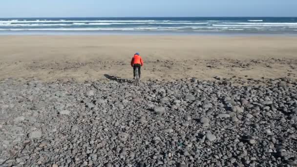 Молодой человек едет на велосипеде по большим камням к песчаному пляжу на Канарских островах. Лароте, Атлантический океан — стоковое видео