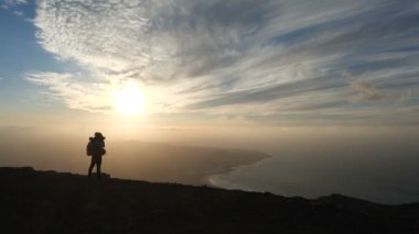 Kanarya Adaları'nda okyanusun üzerinde bir dağın tepesinde inanılmaz bir gün batımı nın fotoğraflarını çeken sırt çantası ile bisiklet kasklı bir kızın silueti. Lanzarote.