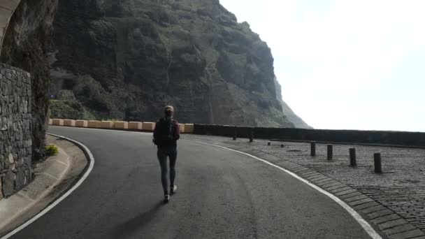 Camera follows young woman with backpack walking from a tunnel towards a cliff edge raising arms in air. — Stock Video