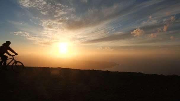 Man rider en cykel hög i bergen nära klippkanten över havet mot vackra dramatiska solnedgång bakgrund. — Stockvideo