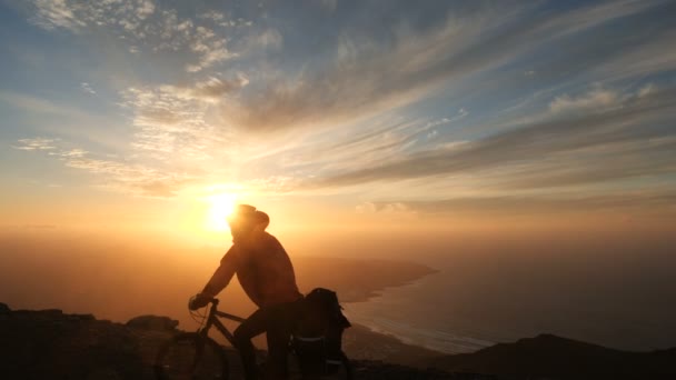 Man rides a bicycle high in mountains near cliff edge above the ocean against beautiful dramatic sunset background. — Stock Video