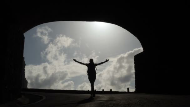 Câmera segue jovem mulher com mochila andando de um túnel em direção a uma borda do penhasco levantando braços no ar . — Vídeo de Stock