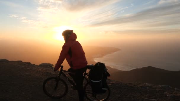 Man on a bicycle high in mountains near cliff edge above the ocean against beautiful dramatic sunset background. — Stock Video