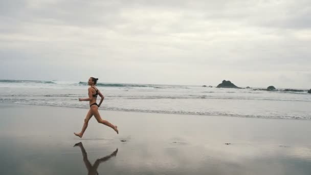 Tracking shot of attractive young woman jogging on stormy Benijo beach in Tenerife, Canary Islands. Female runner in cloudy seaside background — Stock Video