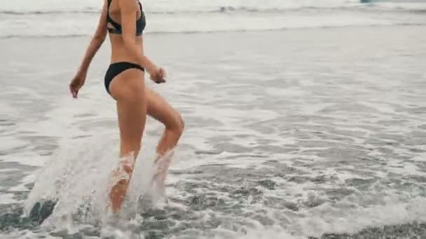 Tracking shot di attraente giovane donna che corre sulla spiaggia di sabbia nera a Tenerife, Isole Canarie. Femmina in nuvoloso sfondo mare — Video Stock