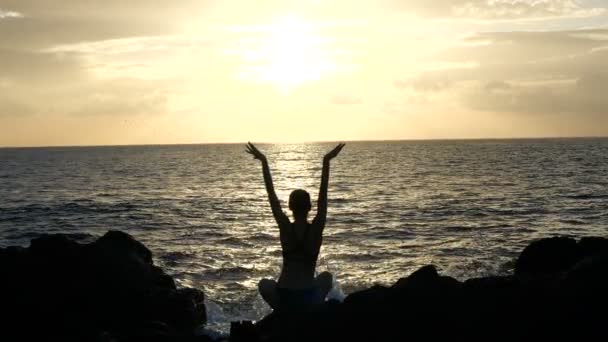 Silhouet van een vrouw zittend op rotsen bij zonsondergang observeren van golven en het verhogen van de armen tegen oceaan golven in filmische Slow Motion — Stockvideo