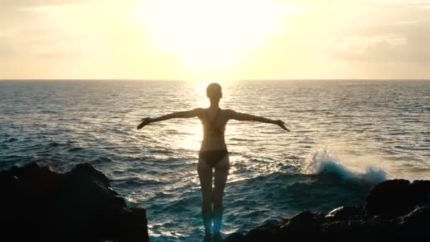 日落时分，一个女人在空中举起双臂，观察巨大的海浪冲击岩石并喷洒。电影慢动作 — 图库视频影像