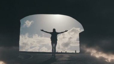 Bir tünelden geçerek havada kollarını kaldıran parlak bir güneş ışığına doğru yürüyen genç bir kadının siluetinin izini sürmek.